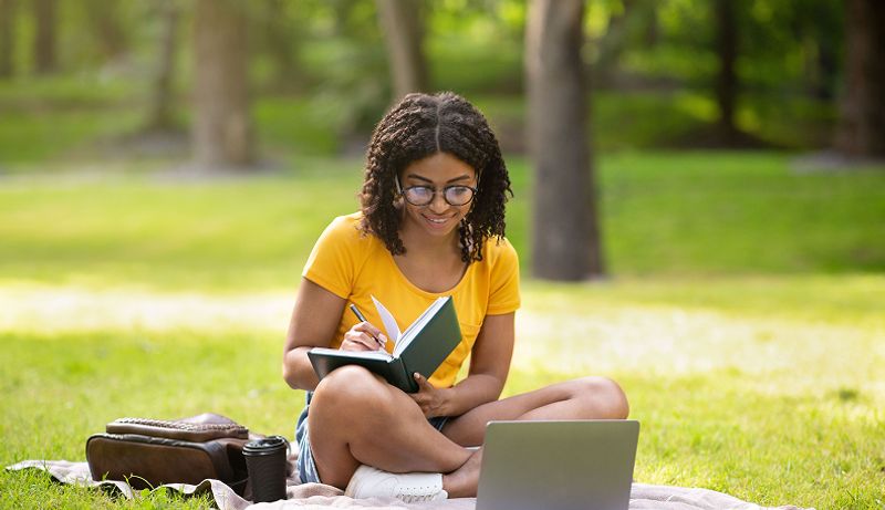 A girl in a yellow writes in a notebook while sitting on the grass
