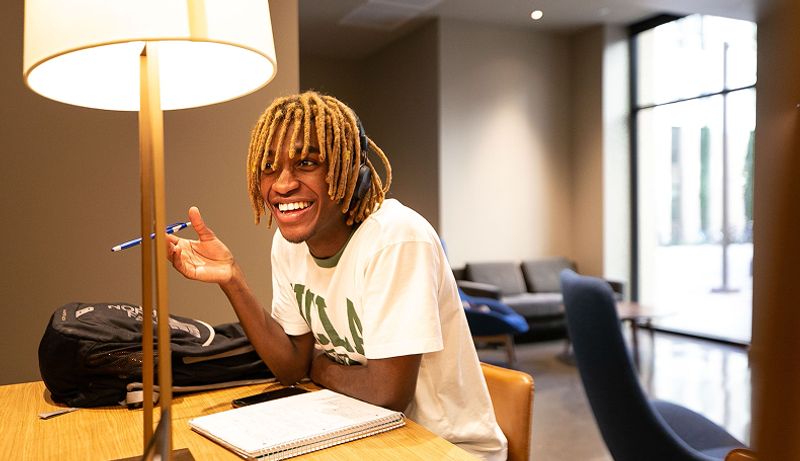 A student wearing headphones and sitting at a desk smiles while looking off camera
