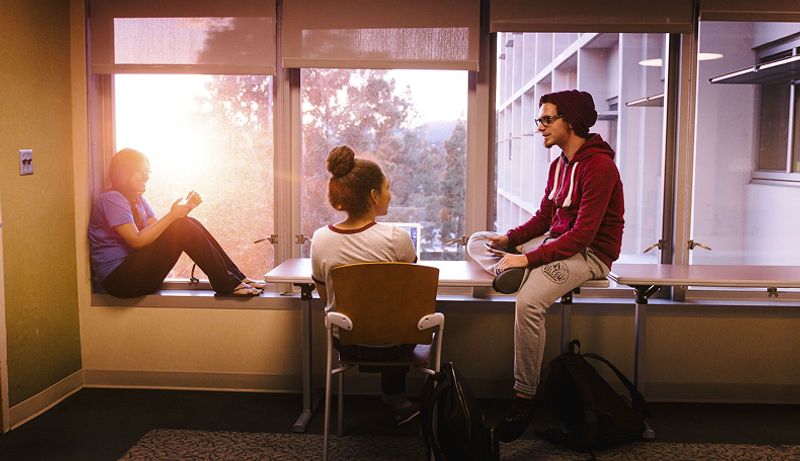 Three students sit and chat by a window. One student sits on the windowsill, one sits on a chair, and one sits on the table facing the others.