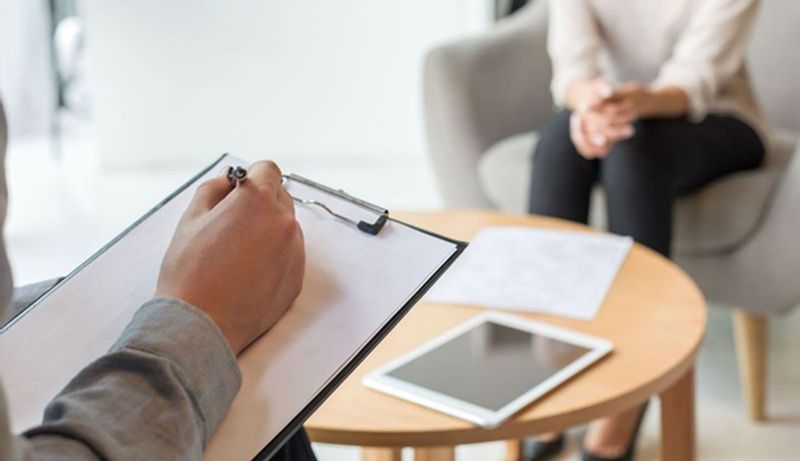 A person writes on a clipboard while a woman sits on a couch behind a table in the background