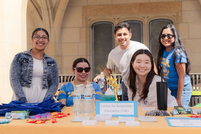Five UCLA Center for Accessible Education representatives smile behind a resource table with informational materials, giveaways, and refreshments at an outdoor campus event