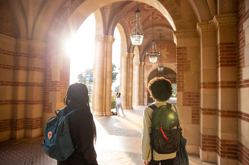 Two students wearing backpacks walk through the sunlit arches of a UCLA campus building