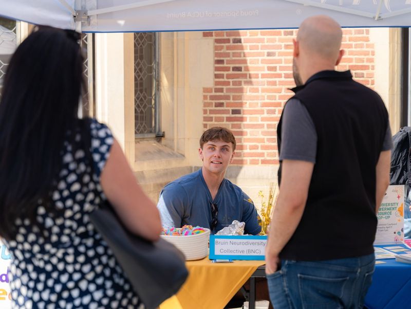 A Bruin Neurodiversity Collective representative speaks with visitors at an outdoor UCLA campus resource fair, seated behind a table displaying informational materials and colorful items
