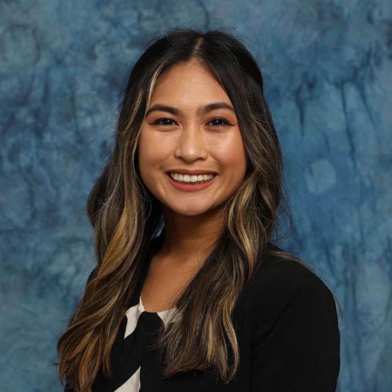 Picture of a Brown woman with brunette and highlighted hair wearing a black shirt in front of a blue background