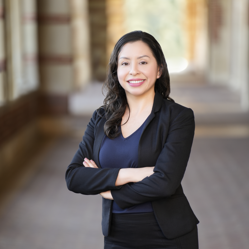 Headshot of white woman with black hair wearing a black suit coat, blue shirt and black trousers